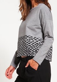 Felpa grigia a maniche lunghe, con un motivo geometrico nero testurizzato nella parte inferiore. Realizzata in tessuto morbido, design corto.