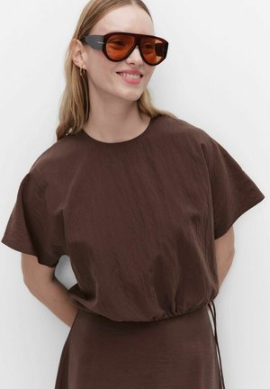 Kvinde smiler iført store brune solbriller, guld øreringe i ringform og en løs brun kortærmet top med samlet talje.