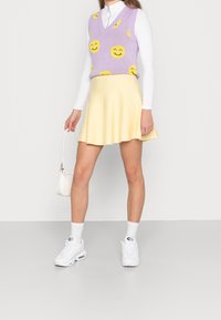 Violet knitted vest with yellow smiley faces, layered over a white mock-neck top. Yellow flared skirt, paired with white sneakers and handbag.