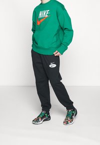 Person som bär en grön Nike-hoodie, svarta Nike-byxor och flerfärgade Nike-sneakers står framför en helt vit bakgrund.