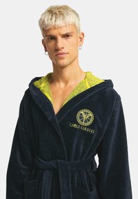 Navyblauwe velours badjas met een capuchon, voorzien van een felgele binnenvoering en geborduurd logo. Inclusief een riem en zijzakken.