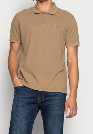 Homme portant un polo beige à manches courtes avec un petit logo rouge Levi's sur la poitrine et un jean bleu, debout avec une main dans la poche.