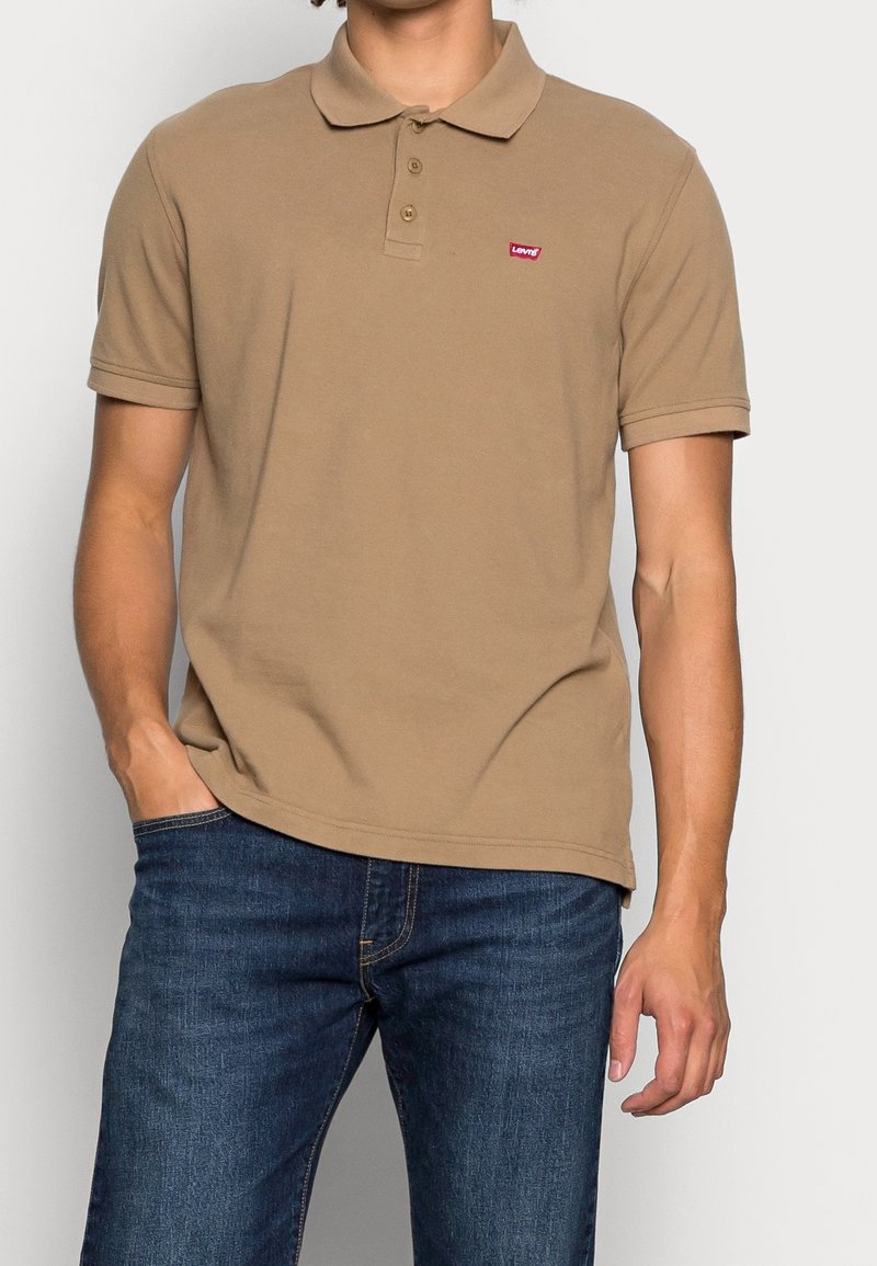 Homme portant un polo beige à manches courtes avec un petit logo rouge Levi's sur la poitrine et un jean bleu, debout avec une main dans la poche.
