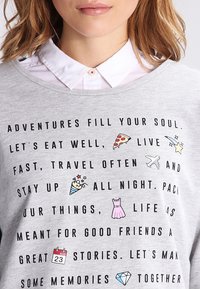 Sudadera gris con cuello redondo, que presenta texto negro y iconos coloridos que representan comida y viajes, sobre un fondo de textura de tela suave.