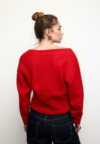 Roter, übergroßer Pullover mit weitem Ausschnitt, langen Ärmeln und geripptem Saum, kombiniert mit dunklen Jeans; glatte Textur, lässiges Design.