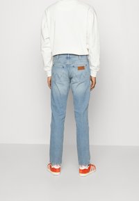 Jeans in denim blu chiaro con taglio dritto e due tasche posteriori. Indossati con una camicia bianca a maniche lunghe e sneaker con dettagli rossi.