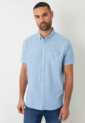 Mann mit kurzen Haaren, der ein hellblaues Kurzarmhemd und blaue Jeans trägt, steht vor einem einfarbigen weißen Hintergrund.