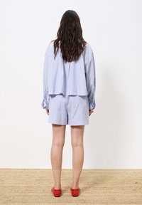 Vrouw staat met haar rug naar de kijker gekeerd en draagt een lichtblauw gestreept shirt met lange mouwen en shorts, met rode flatschoenen op een geweven tapijt.