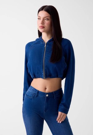 Frau mit langen dunklen Haaren, die ein blaues, bauchfreies Zip-Hoodie und eine hoch taillierte blaue Jeans trägt, steht vor einem schlichten hellen Hintergrund.