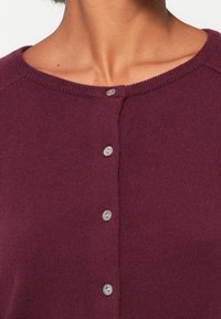 Cárdigan de punto burdeos con cuello redondo y cinco botones de color claro en la parte delantera, con una textura suave y lisa.