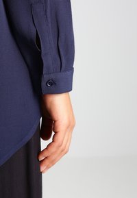 Marinblå skjorta med textilstruktur, med långa ärmar och knappade manschetter. Sömmen är lätt rundad. Närbild på hand på ärmen.