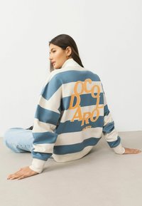 Striped sweatshirt in blue and cream with thick horizontal lines. Features orange text on the back. Slightly oversized fit.