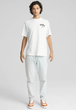 Witte katoenen t-shirt met korte mouwen, voorzien van een zwart Puma-logo. Gecombineerd met lichtblauwe jeans en sneakers met oranje accenten.