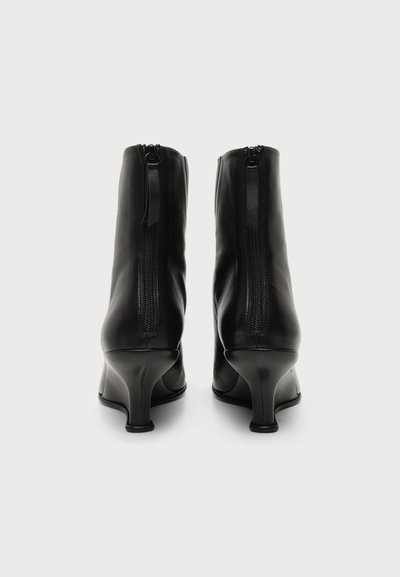 Paire de bottines en cuir noir avec zips arrière et talons bas incurvés, vues de dos sur un fond uni.