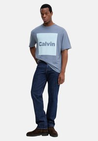 Camiseta de algodón gris con un gran cuadrado azul claro que muestra la palabra "Calvin" en azul oscuro, combinada con jeans de mezclilla oscuros y botas marrones.