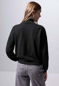 Maglione nero in maglia con colletto alto, vestibilità comoda e orlo a costine, abbinato a jeans in denim grigio chiaro. Texture morbida, design minimal.