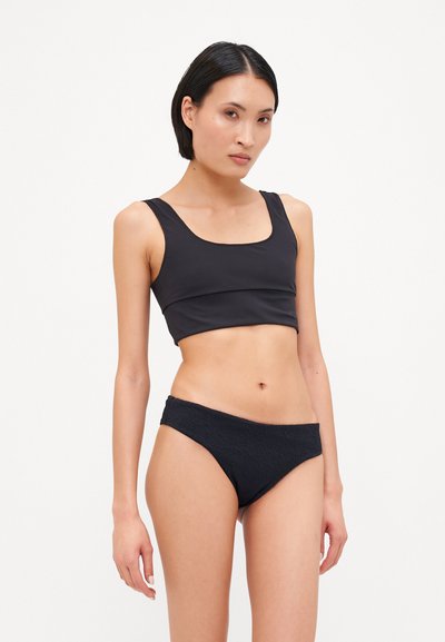 Melna bikini komplekts ar cieši pieguļošu, izgrieztu augšdaļu un teksturētām zemo vidukļa biksītēm, abas izgatavotas no gludas, elastīgas materiāla.