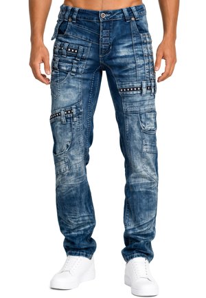 Man wearing blue denim cargo jeans with multiple pockets, black detailing, and white sneakers, standing against a white background.