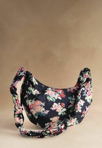 Mustast poolkuukujulisest käekotist, millel on roosa ja roheline lillemuster ning kokkutõmbevõlviga rihm, beeži tausta ees.