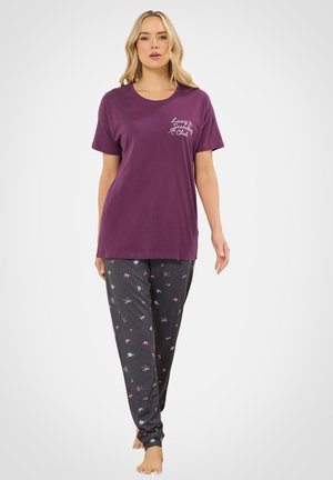 Kobieta w fioletowej koszulce z krótkim rękawem z napisem "Lazy Sunday Club" oraz ciemnych spodniach do piżamy z małymi kolorowymi wzorami, stojąca boso.