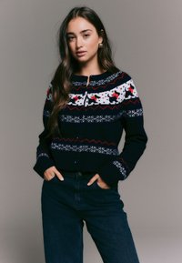 Tumesinine kardigan valgete ja punaste pidulike mustritega, ümmarguse kaelusega, nööpidega kinnitatav, soonikkoes varrukaservade ja lühendatud äärega, stiilitud kõrge vöökohaga teksadega.