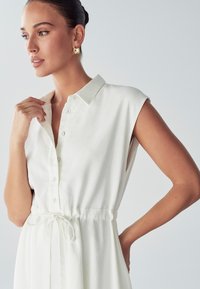 Vestido blanco con cuello camisero, mangas cortas, cierre de botones y cintura ajustable. Tejido suave con un ligero brillo y corte relajado.