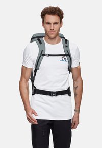 Grå ryggsäck med justerbara svarta remmar och spännen, vadderad baksida och en logotyp. Bärs över en vit t-shirt, tillsammans med svarta byxor.