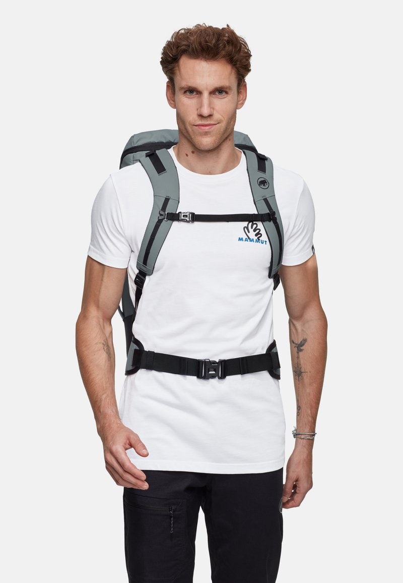 Grå ryggsäck med justerbara svarta remmar och spännen, vadderad baksida och en logotyp. Bärs över en vit t-shirt, tillsammans med svarta byxor.
