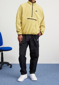 Gul fleecepullover med halvdragkedja och svart dragkedjeficka, kombinerad med svarta cargobyxor och vita sneakers.