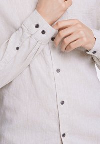 Camisa de lino beige con acabado texturizado, que presenta botones negros en los puños y la tapeta, y un único bolsillo en el pecho.