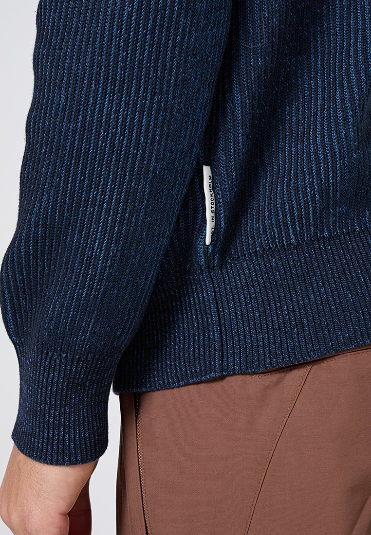 Navyblaues Strickpulloverärmel und Seitennähte mit weißem "Made in Stockholm"-Aufnäher, über braunen Hosen, Handgelenk und Hand sichtbar.