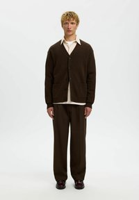 Brauner gestrickter Cardigan mit großen Knöpfen und seitlichen Taschen, über einem hellen Hemd getragen. Kombiniert mit braunen Hosen und dunklen Schuhen.