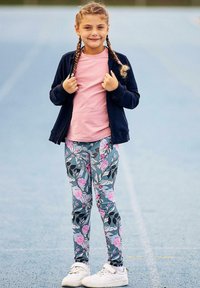 Młoda dziewczyna z warkoczami, ubrana w różową koszulkę, kwiatowe legginsy, granatową kurtkę i białe sneakersy, stoi na niebieskim torze sportowym.