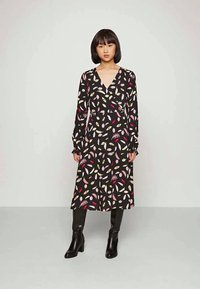 Vestido preto de manga longa com um design envolvente, apresentando um padrão colorido de folhas em rosa, amarelo e branco. Usado com botas pretas até ao joelho.