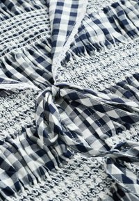 Tissu à carreaux bleu et blanc avec des sections en dentelle blanche smockée et un détail de nœud noué au centre.