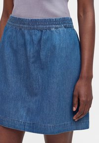 Femme portant une jupe en jean bleue longueur genou avec une ceinture élastique et un haut violet clair rentré à l’intérieur, debout avec la main le long du corps.