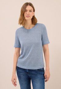 Eine Frau trägt ein leicht blaues, kurzärmliges Strickoberteil, das sie mit mittelblauen Jeans kombiniert hat, während sie vor einem schlichten beigen Hintergrund steht.