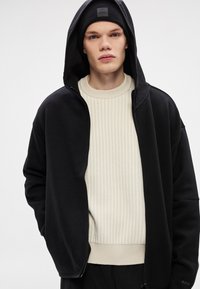 Μαύρο φούτερ με φερμουάρ και επένδυση με υφή, φορεμένο πάνω από ένα ανοιχτό μπεζ πουλόβερ με ραβδώσεις. Το μοντέλο φορά επίσης μαύρο σκούφο με λογότυπο.