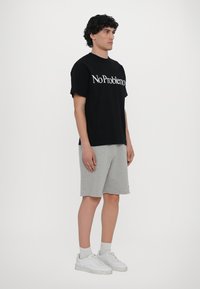 Svart bomulls T-skjorte med "No Problemo" i hvit tekst, kombinert med grå shorts og hvite sneakers. Enkelt design, avslappet passform, standard hals.