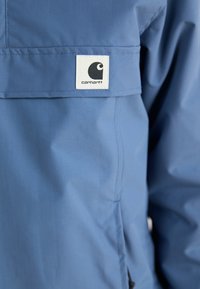 Giacca azzurra chiara con una texture liscia, dotata di una toppa con logo bianco. Il design include una tasca frontale con patta e dettagli minimalisti.
