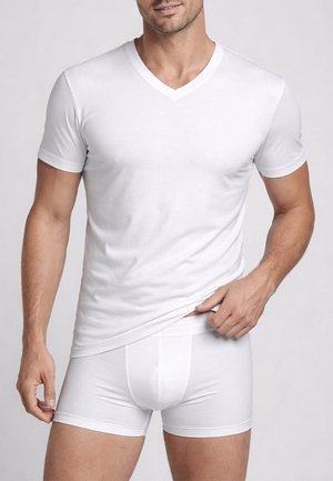 Uomo che indossa una T-shirt bianca a scollo a V e boxer bianchi, che aggiusta il bordo inferiore della T-shirt con una mano.