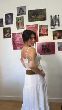 Γυναίκα με λευκό τοπ και φούστα με τατουάζ στο χέρι στέκεται πλάγια μπροστά σε έναν τοίχο διακοσμημένο με διάφορα έργα τέχνης και φωτογραφίες.