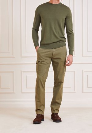Homme portant un pull à manches longues vert olive, un pantalon cargo kaki avec poches latérales, et des bottes à lacets marron foncé, debout à l'intérieur.