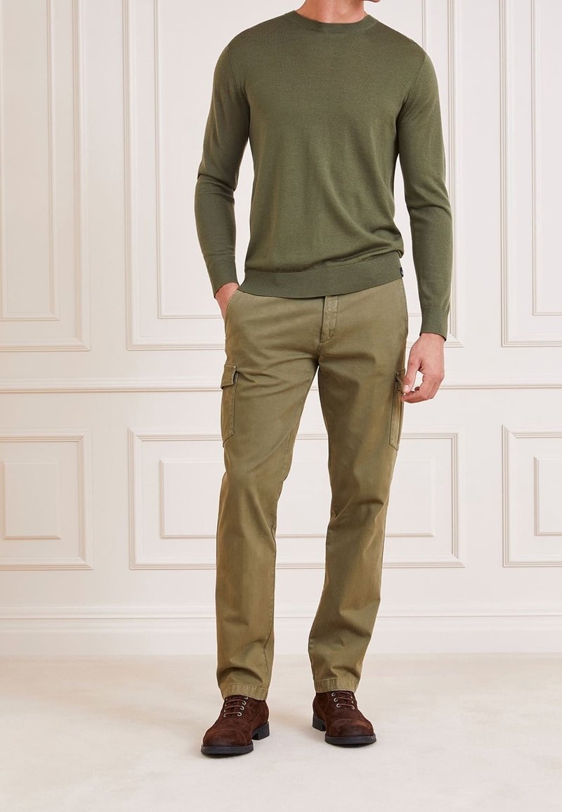 Homme portant un pull à manches longues vert olive, un pantalon cargo kaki avec poches latérales, et des bottes à lacets marron foncé, debout à l'intérieur.