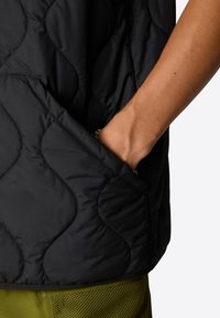 Black quilted vest with curved stitching, featuring a side pocket. The texture is smooth, complemented by a green mesh base layer.