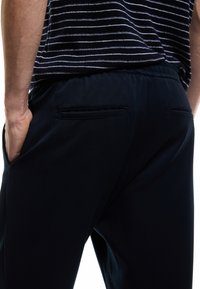 Dunkelblaue Hose mit glatter Textur, elastischem Bund, zwei Gesäßtaschen und subtilen Schnittdetails, kombiniert mit einem gestreiften Hemd.