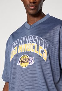Grå mesh baskettröja med v-ringad hals, med "LOS ANGELES" i vitt och guld, samt "LAKERS"-logotypen i lila och gult.