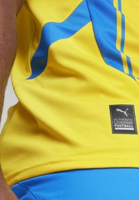 Gul fodboldtrøje med blå detaljer og en struktureret stof. Har et sort label med teksten "AUTENTISK LICENSED FODBOLD" fra Puma.