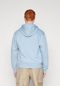 Ljusblå hoodie i mjukt material, med dragsko i huvan och ribbade ärmslut. Kombinerad med tanfärgade cargobyxor, visad bakifrån.