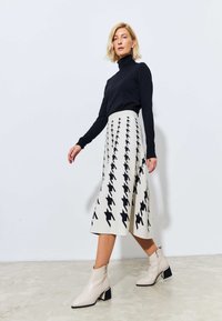 Un pulover negru cu guler înalt, asociat cu o fustă crem cu un model de houndstooth negru, completat de cizme scurte bej cu toc pătrat.